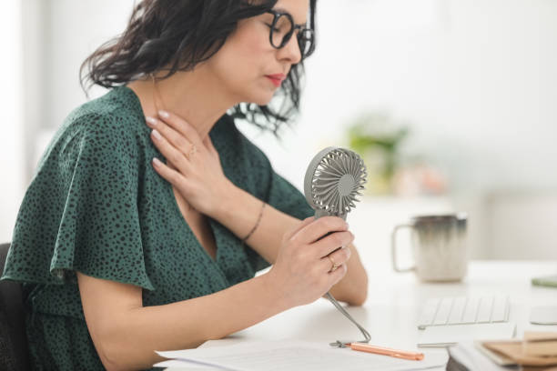 Woman in her 40s using a portable desk fan at work to cool down during a menopause hot flash 